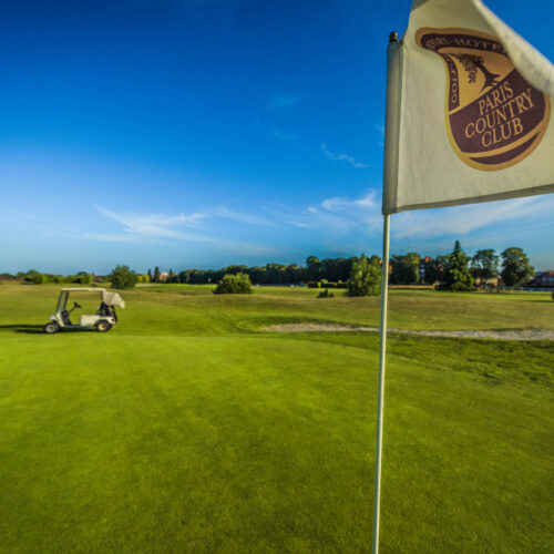 Drapeau du Paris Country Club au milieu d'un green. Caddy au fond, zone de sable sur la droite